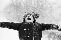 A person wearing a dark winter coat and a fuzzy pom-pom hat throws their arms wide in the snow, with snowflakes falling around them in a black and white outdoor scene.