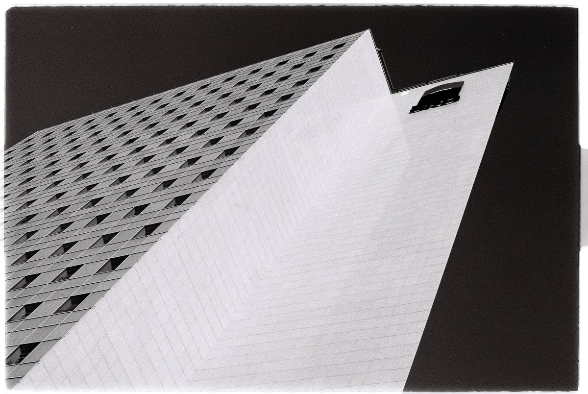 Black and white architectural photograph of a modernist building with dramatic perspective angles, featuring a grid-patterned facade and a large white corner plane against a dark sky.