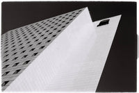 Black and white architectural photograph of a modernist building with dramatic perspective angles, featuring a grid-patterned facade and a large white corner plane against a dark sky.
