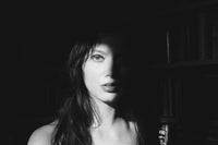 Black and white portrait of a woman with long dark hair, shown in dramatic side lighting against a dark background with bookshelves visible.