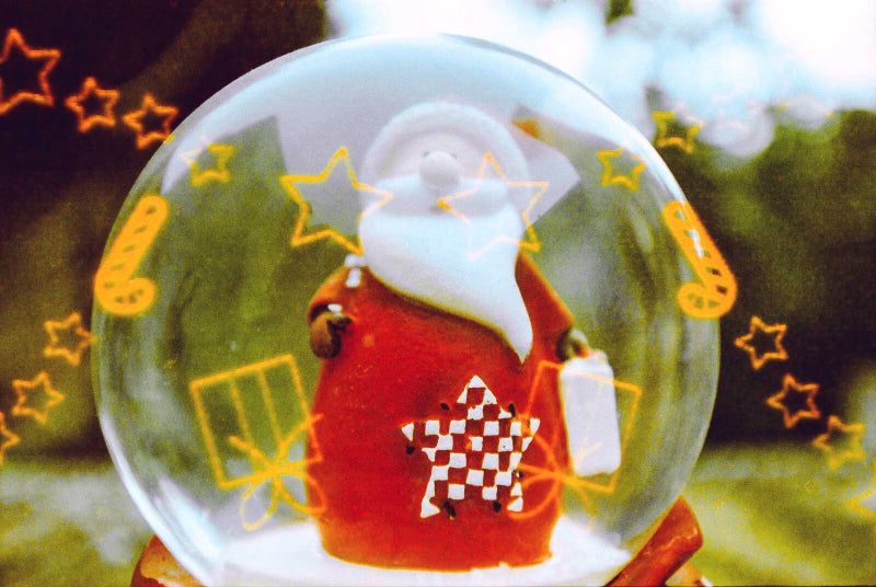 A clear glass snow globe featuring a small Santa Claus figure in a red suit with a white beard, surrounded by yellow star and gift graphics, set against a blurred outdoor background.