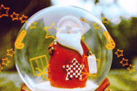 A clear glass snow globe featuring a small Santa Claus figure in a red suit with a white beard, surrounded by yellow star and gift graphics, set against a blurred outdoor background.