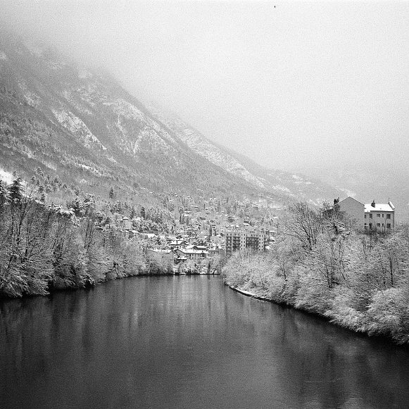Black and white landscape photograph of a snowy mountain valley with a river in the foreground, snow-covered trees, a small town, and a building on the right.