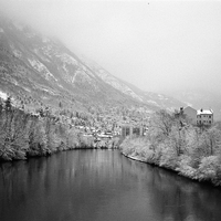 Black and white landscape photograph of a snowy mountain valley with a river in the foreground, snow-covered trees, a small town, and a building on the right.
