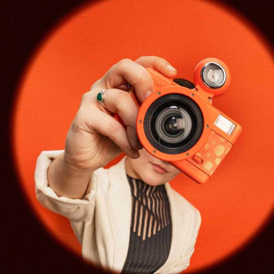 A person holding an orange compact camera up to their face, framed by a circular orange spotlight against a dark background.