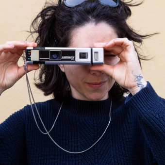 A person holding a vintage Leica SL camera to their eye, wearing a dark navy knit sweater against a plain beige background.