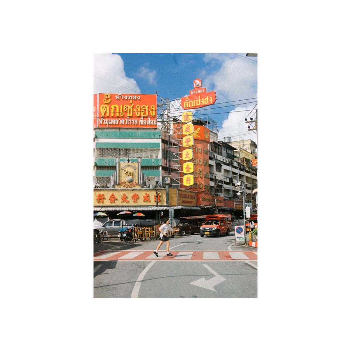A vertical rectangular photo print or poster showing a street scene in Bangkok with colorful orange and yellow shop signs, cars, a crosswalk, and a pedestrian under a blue sky with clouds, centered on a white background.