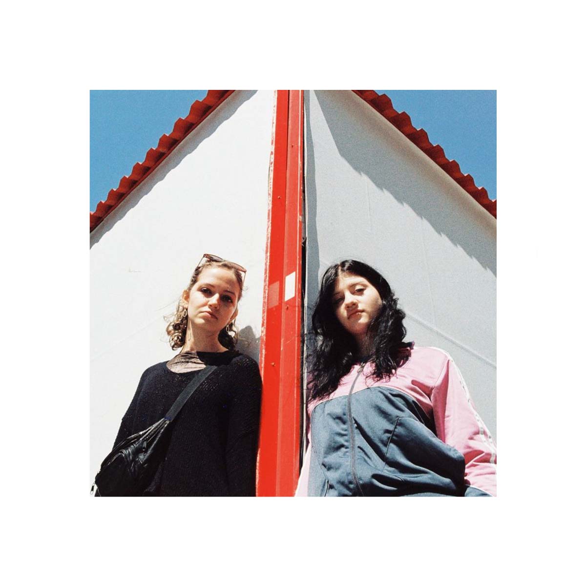 Two women posing against a white building with a red roof ridge and blue sky, styled in a minimalist, editorial fashion.