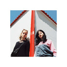 Two women posing against a white building with a red roof ridge and blue sky, styled in a minimalist, editorial fashion.