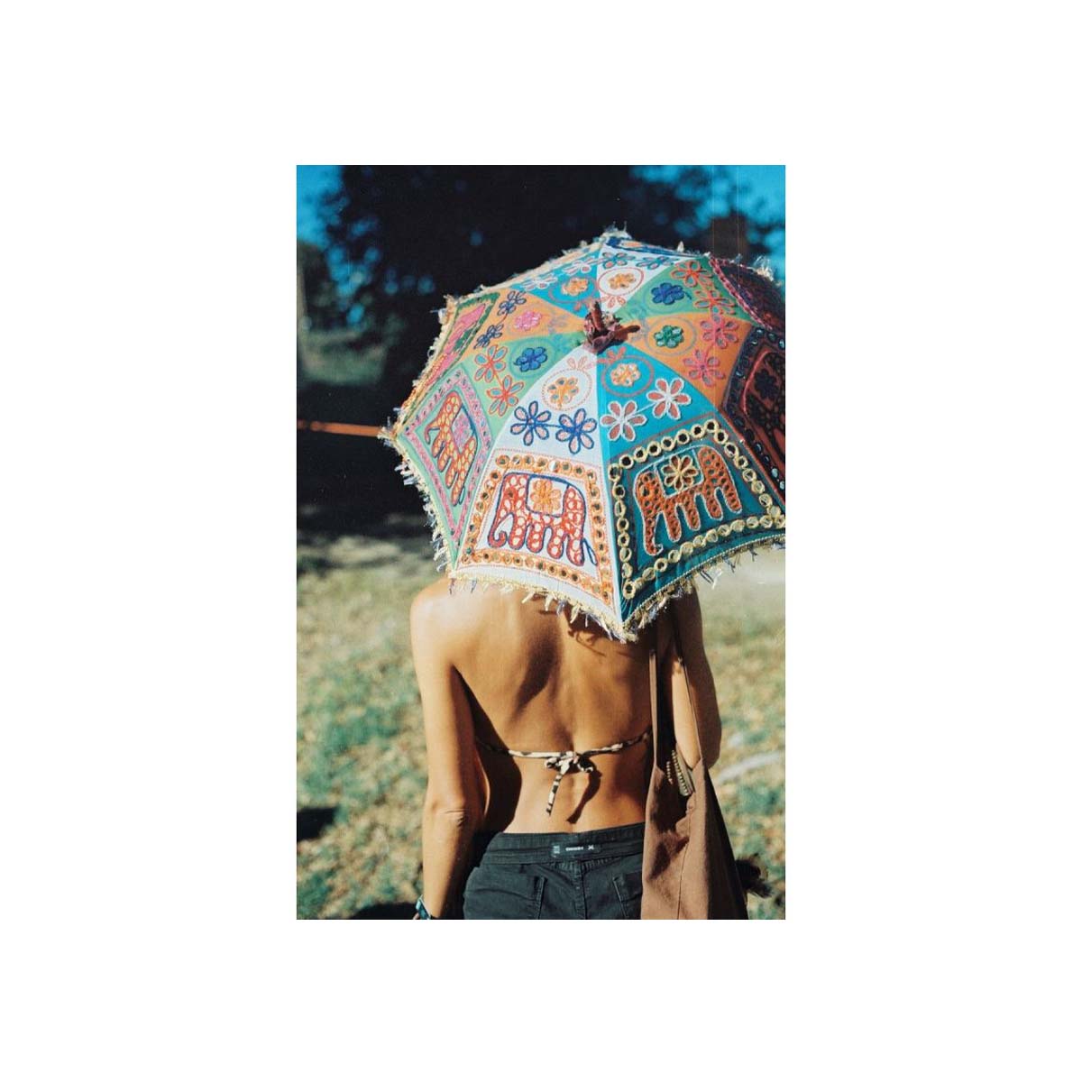 A woman seen from behind holding a large, colorful bohemian-style parasol umbrella outdoors. The umbrella features intricate patterns with elephant motifs and fringed edges.