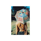A woman seen from behind holding a large, colorful bohemian-style parasol umbrella outdoors. The umbrella features intricate patterns with elephant motifs and fringed edges.