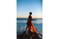 Silhouette of a person in a flowing orange dress walking through ocean waves at sunset, shot on LomoChrome Color '92 film.