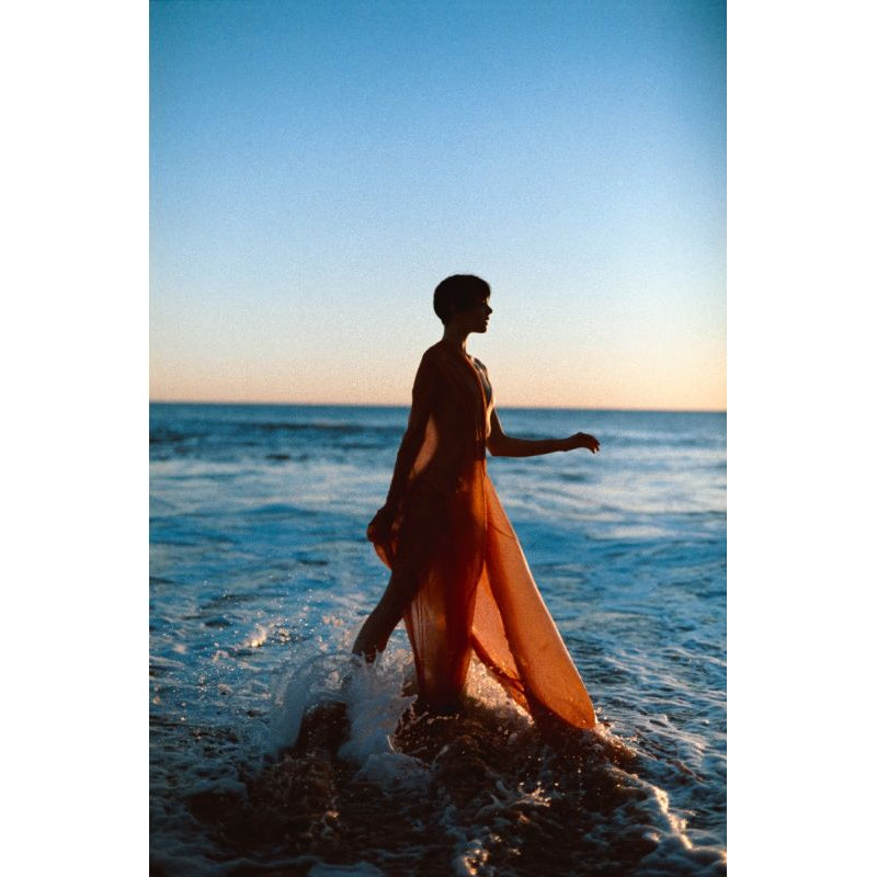 Silhouette of a person in a flowing orange dress walking through ocean waves at sunset, shot on LomoChrome Color '92 film.