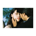 A close-up photograph of a woman with long dark hair being gently touched by a man in a black suit outdoors. The photo has a soft, slightly filtered vintage aesthetic with blue sky and foliage in the background.