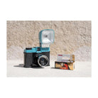 A retro-style instant camera with a turquoise and black body and an attached flash, shown next to a box of Colorplus Color ISO 600 film on a light concrete surface against a textured beige wall.