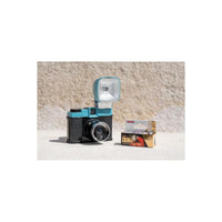 A retro-style instant camera with a turquoise and black body and an attached flash, shown next to a box of Colorplus Color ISO 600 film on a light concrete surface against a textured beige wall.