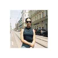 A woman wearing a black sleeveless mock-neck top and sunglasses stands on a city street with buildings and parked cars in the background.