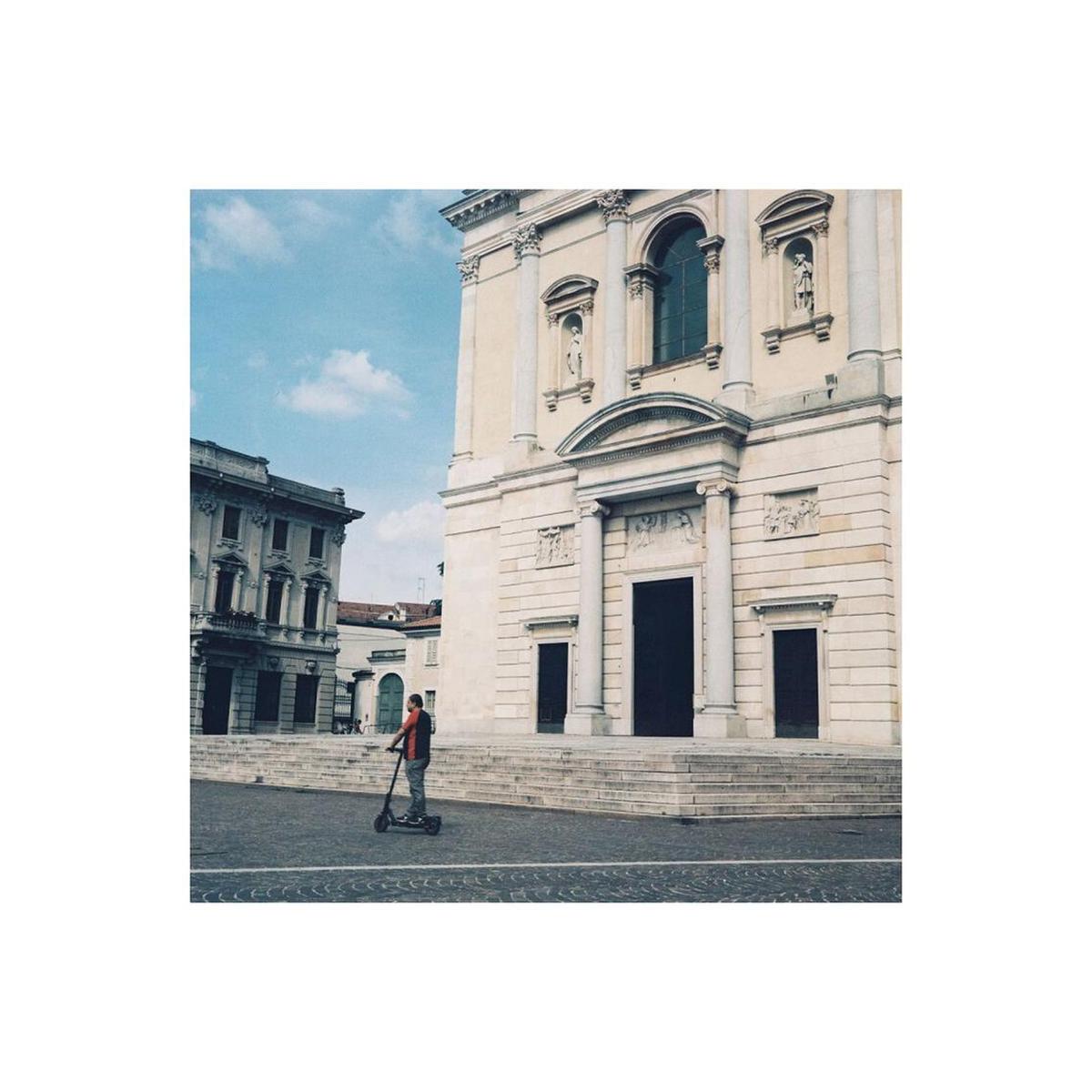 A person riding a black electric scooter in a city square in front of a large beige stone building with classical architecture under a blue sky.