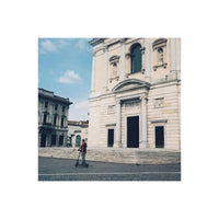 A person riding a black electric scooter in a city square in front of a large beige stone building with classical architecture under a blue sky.