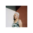 An older woman with short white hair stands against a split white and reddish-brown wall, wearing a green and white patterned sleeveless top and large round earrings.