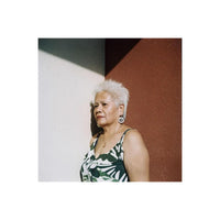 An older woman with short white hair stands against a split white and reddish-brown wall, wearing a green and white patterned sleeveless top and large round earrings.