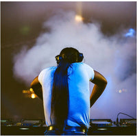 A person seen from behind wearing a white t-shirt and headphones, standing at DJ equipment with smoke and blue stage lighting in the background.