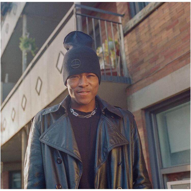 A man wearing a black knit beanie with a small circular logo, a black leather jacket, and a silver chain necklace, standing outdoors in front of a brick building.