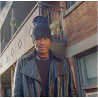 A man wearing a black knit beanie with a small circular logo, a black leather jacket, and a silver chain necklace, standing outdoors in front of a brick building.