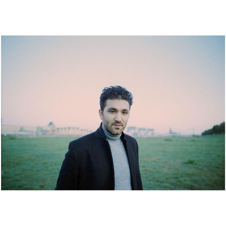 A man with dark curly hair and a light beard, wearing a black coat over a gray turtleneck sweater, standing in an open grassy field under a pale pink and blue sky.