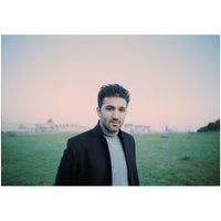 A man with dark curly hair and a light beard, wearing a black coat over a gray turtleneck sweater, standing in an open grassy field under a pale pink and blue sky.
