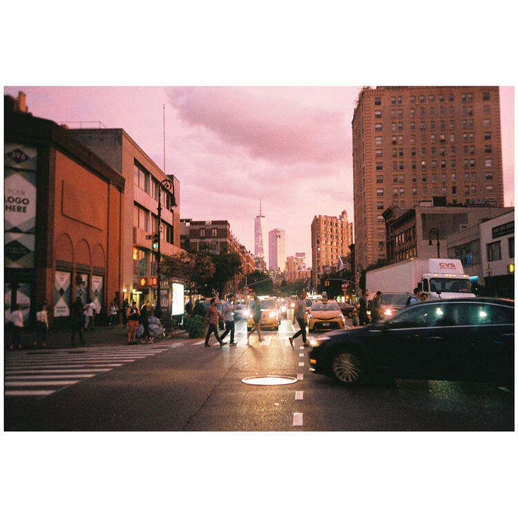 A vintage-style photograph of a city street at dusk, featuring pedestrians crossing at a crosswalk, cars on the road, and tall buildings under a pinkish sky. The image is presented with white letterbox bars at the top and bottom.