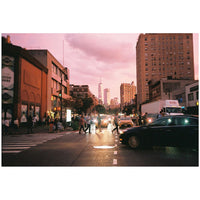 A vintage-style photograph of a city street at dusk, featuring pedestrians crossing at a crosswalk, cars on the road, and tall buildings under a pinkish sky. The image is presented with white letterbox bars at the top and bottom.