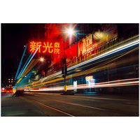 A long-exposure city street scene at night with bright red and yellow neon signage, blurred vehicle light trails, and a dark urban background.