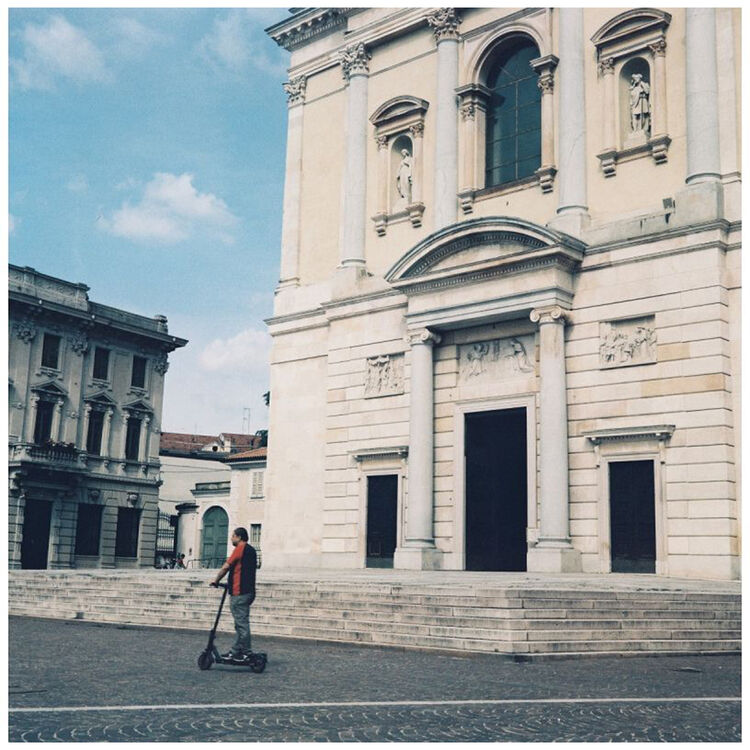 A person riding a black electric scooter in a city square in front of a large beige stone building with classical architecture under a blue sky.