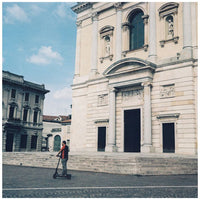 A person riding a black electric scooter in a city square in front of a large beige stone building with classical architecture under a blue sky.