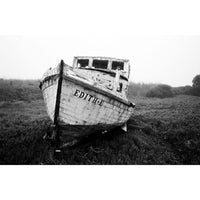 A black and white photograph of an old, weathered wooden boat labeled 'EDITH' resting on a trailer in an open grassy field.
