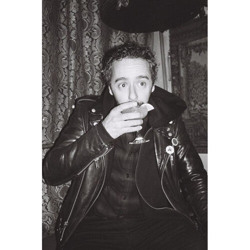 A black and white portrait of a man wearing a shiny leather jacket, holding a glass near his mouth, with patterned curtains and a dark hat in the background.