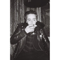 A black and white portrait of a man wearing a shiny leather jacket, holding a glass near his mouth, with patterned curtains and a dark hat in the background.