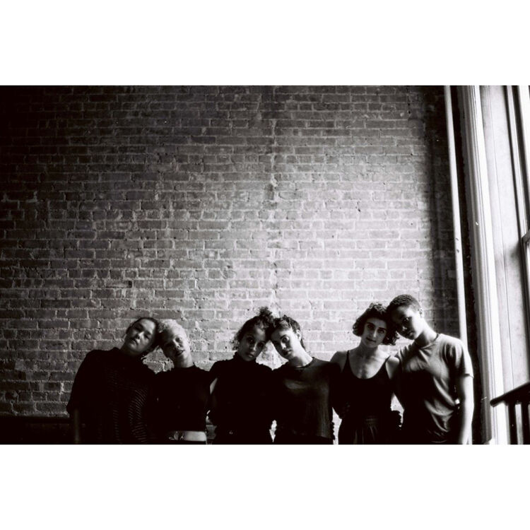 Black and white photograph of five people standing together against a brick wall, with a window light shining in from the right side.