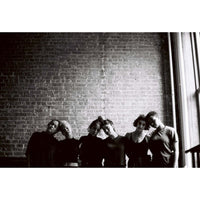 Black and white photograph of five people standing together against a brick wall, with a window light shining in from the right side.