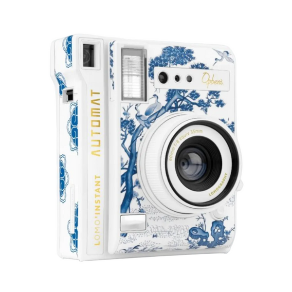 A white instant camera with a blue porcelain-style floral and bird pattern, featuring gold lettering that reads 'LOMOINSTANT AUTOMAT' and 'Ophiri'.
