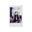 A square instant photograph print with a white border, featuring a woman holding a small camera in front of her face, set against a plain white background.