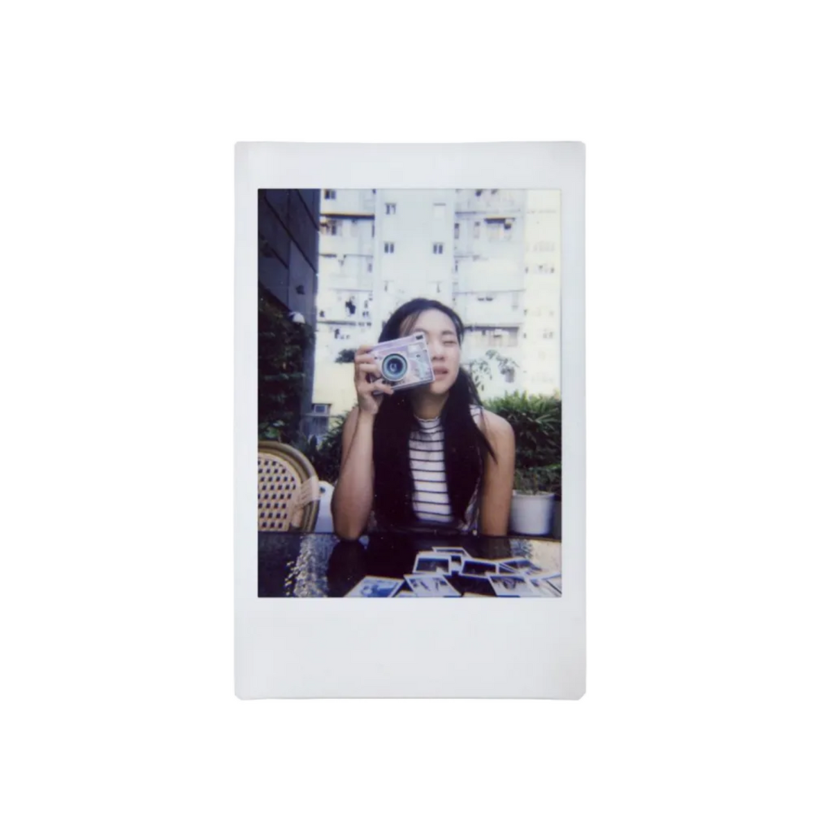 A square instant photograph print with a white border, featuring a woman holding a small camera in front of her face, set against a plain white background.