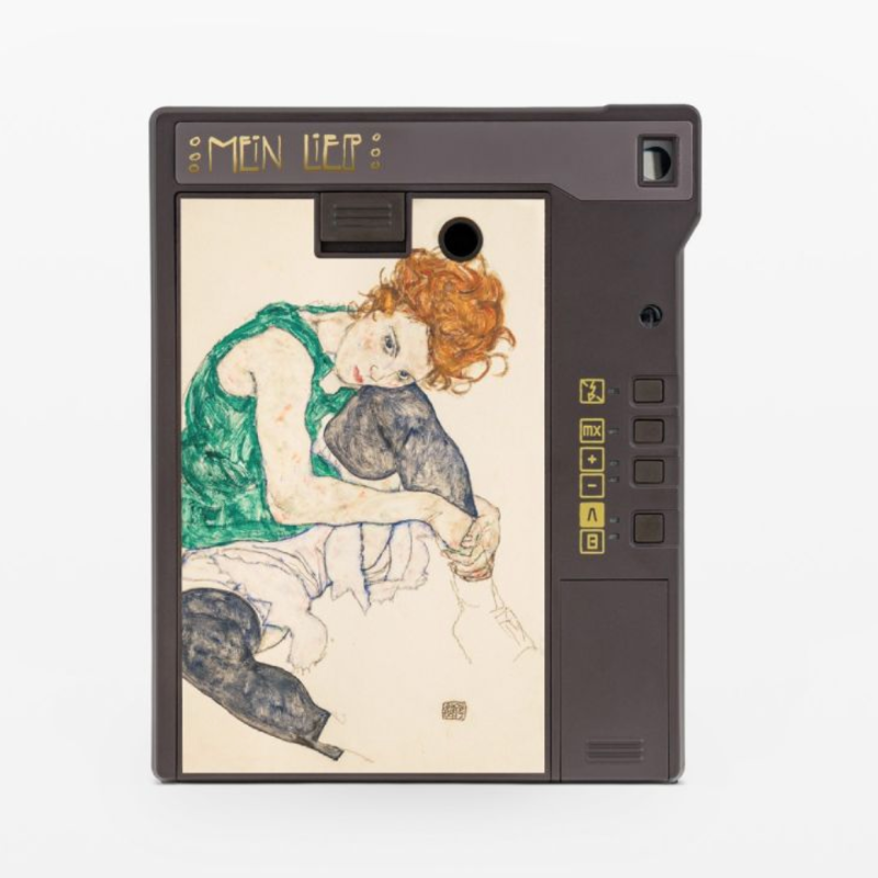 A retro-style compact camera with a dark gray body and a screen displaying an Art Nouveau illustration of a woman with red hair. The camera has gold lettering that reads 'MEN & LIFE' on the top left, a small lens window on the top right, and a vertical row of yellow icons on the right side.