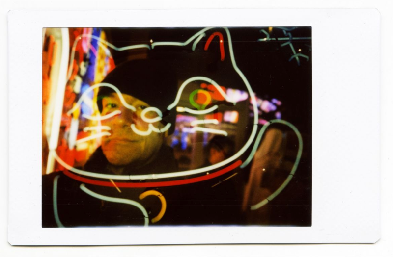A square instant photograph with a white border showing a neon-lit cat-shaped display at night, with a blurred face reflected in the glass.