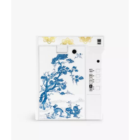 A white instant camera featuring a blue and white porcelain-style floral and bird illustration on the back panel, with gold decorative accents near the top and a white side control panel.