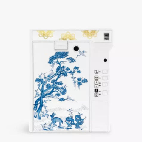 A white instant camera featuring a blue and white porcelain-style floral and bird illustration on the back panel, with gold decorative accents near the top and a white side control panel.