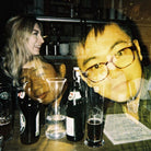 A dimly lit bar scene with several people visible through a reflective glass surface, along with beer bottles, glasses, and a menu on the counter.