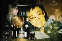 A dimly lit bar scene with several people visible through a reflective glass surface, along with beer bottles, glasses, and a menu on the counter.