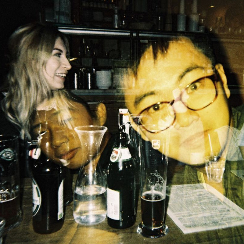 A dimly lit bar scene with several people visible through a reflective glass surface, along with beer bottles, glasses, and a menu on the counter.